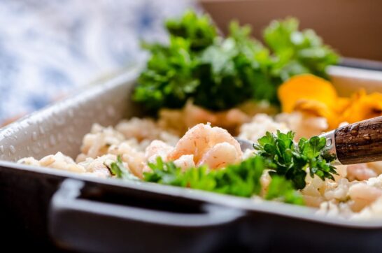 Uma deliciosa porção de risoto de camarão, servido em um prato branco decorado com camarões suculentos, ervas frescas e um toque de crocante. O prato é um verdadeiro festim visual, com os camarões dourados reluzindo sobre o arroz cremoso, adornado com folhas de agrião e rúcula fresca. Uma iguaria irresistível para os amantes de frutos do mar e gastronomia refinada